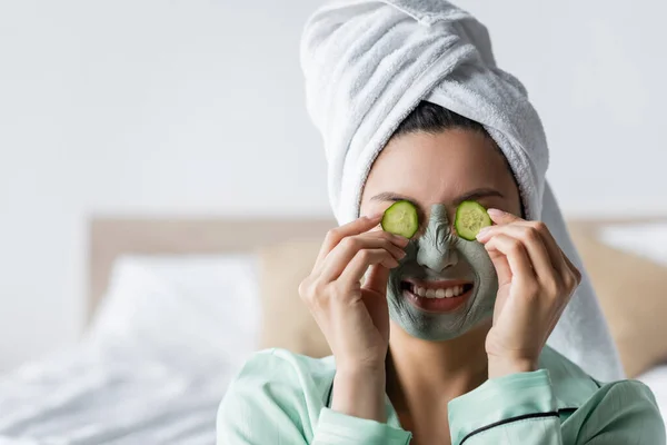 Cheerful asian woman in clay mask frowning while applying cucumber slices on eyes — Stock Photo
