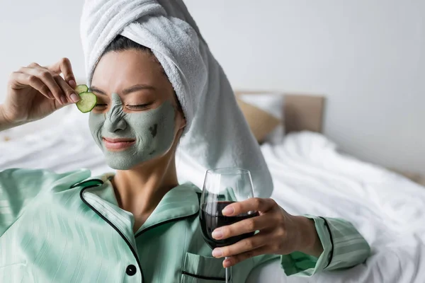 Cheerful sian woman in clay mask applying cucumber slices on eye while holding glass of red wine — Stock Photo