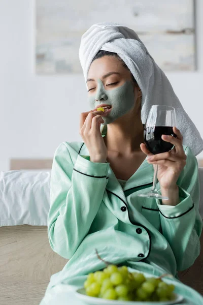 Asiatin in Handtuch auf dem Kopf und Lehmmaske, die Weintrauben isst, während sie ein Glas Rotwein in der Hand hält — Stockfoto