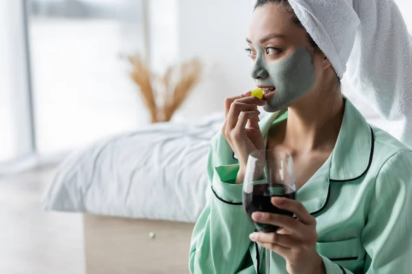 Asiatico donna in argilla maschera mangiare uva mentre holding bicchiere di rosso vino in camera da letto — Foto stock