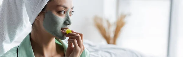 Young asian woman in clay mask and towel on head eating grape in bedroom, banner — Stock Photo