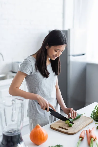 Feliz asiático mujer corte pepino cerca fresco zanahorias y calabaza en cocina - foto de stock