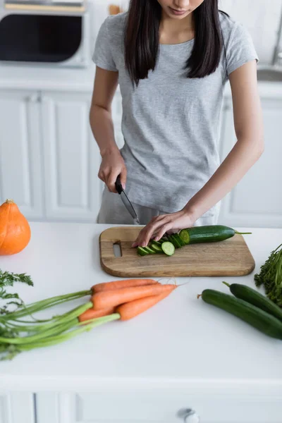 Teilansicht einer Frau beim Gurkenschneiden in der Nähe von frischen Möhren und Kürbis in der Küche — Stockfoto