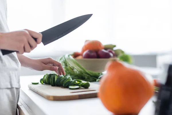 Partial view of woman with knife near sliced cucumber on chopping board and pumpkin on blurred foreground — Stock Photo