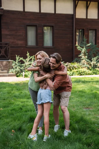 Sorrindo pais abraçando criança na grama perto de casa — Fotografia de Stock