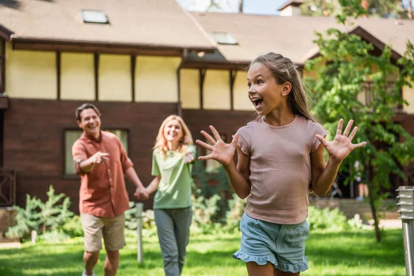 Criança positiva de pé perto dos pais e casa de férias turva — Fotografia de Stock