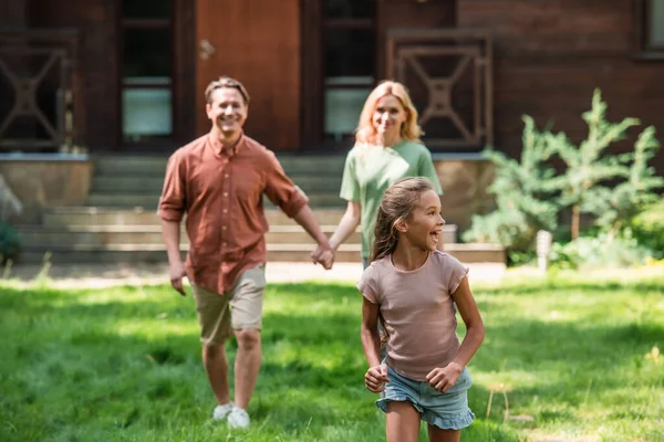 Happy child walking on lawn near blurred parents during weekend — Stock Photo