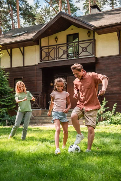 Smiling man playing football with daughter near wife with smartphone — Stock Photo