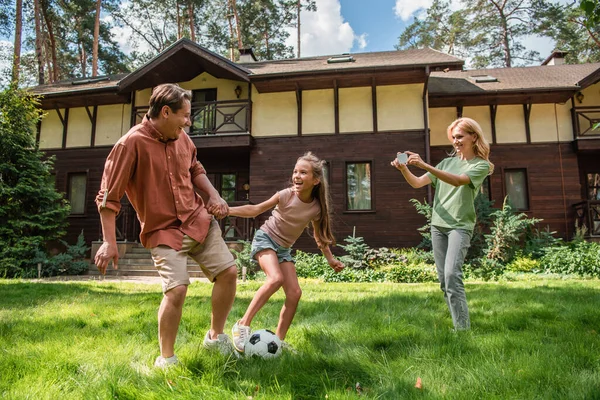 Padre e hijo jugando al fútbol cerca de la madre con teléfono inteligente en el césped - foto de stock