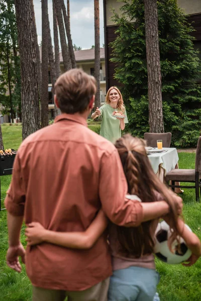 Улыбающаяся женщина держит еду и указывает на размытую семью с футбольным мячом на открытом воздухе — стоковое фото
