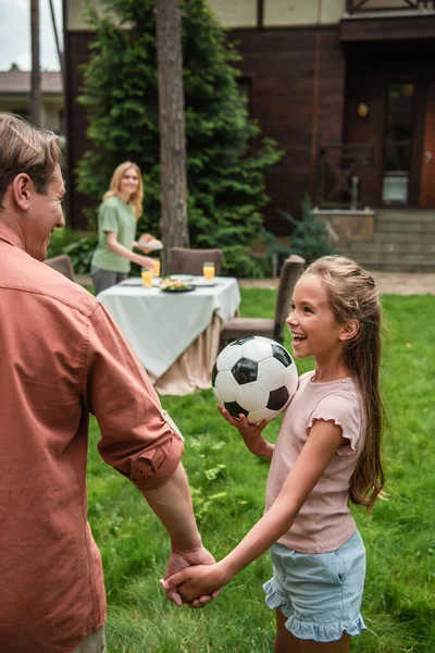 Sonriente niño sosteniendo pelota de fútbol y sosteniendo la mano del padre al aire libre - foto de stock