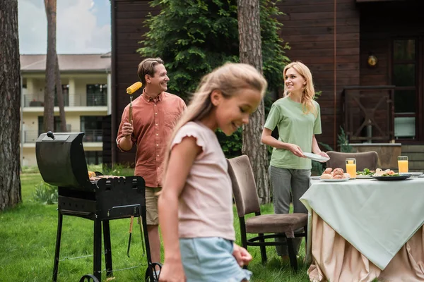 Smiling man baking corn near wife and smiling kid on lawn — Stock Photo