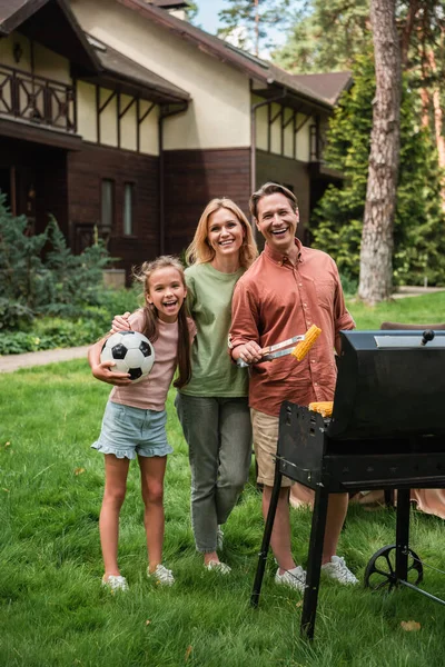 Familia sonriente con pelota de fútbol y maíz de pie cerca de la parrilla al aire libre - foto de stock