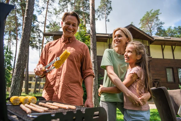 Happy man holding corn near grill and family outdoors — Stock Photo