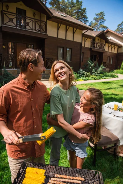 Man embracing smiling family while preparing corn on grill outdoors — Stock Photo