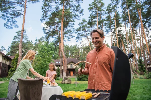 Lächelnder Mann mit Wurst am Grill und verschwommener Familie im Freien — Stockfoto