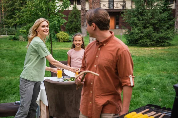 Lächelnde Mutter und Kind schauen Vater in der Nähe des verschwommenen Grills im Freien an — Stockfoto