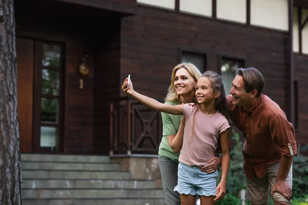 Ragazza sorridente che prende selfie con i genitori vicino alla casa di vacanza offuscata — Foto stock