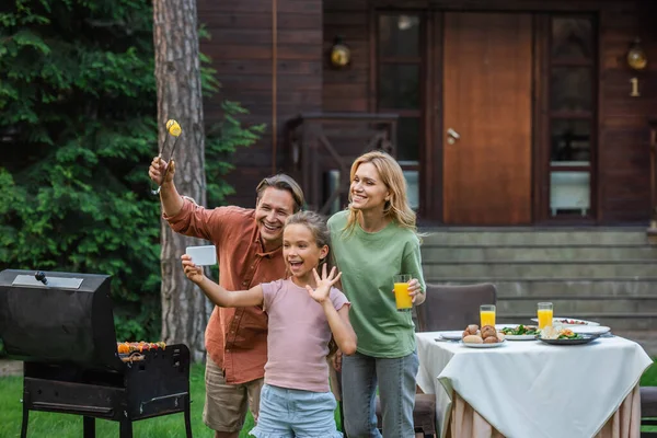 Familia alegre con comida y jugo de naranja teniendo videollamada cerca de la parrilla al aire libre - foto de stock