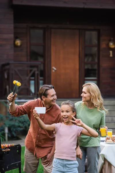 Enfant heureux montrant signe de paix tout en prenant selfie près des parents avec de la nourriture à l'extérieur — Photo de stock
