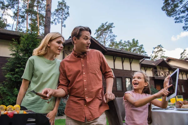 Smiling kid pointing at digital tablet near parents and grill outdoors — Stock Photo