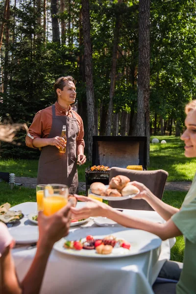 Man in apron holding bottle of beer near blurred family with food outdoors — Stock Photo