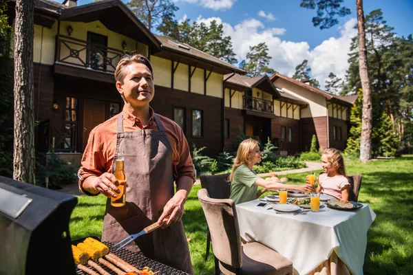 Man holding beer while cooking on grill near smiling family outdoors — Stock Photo