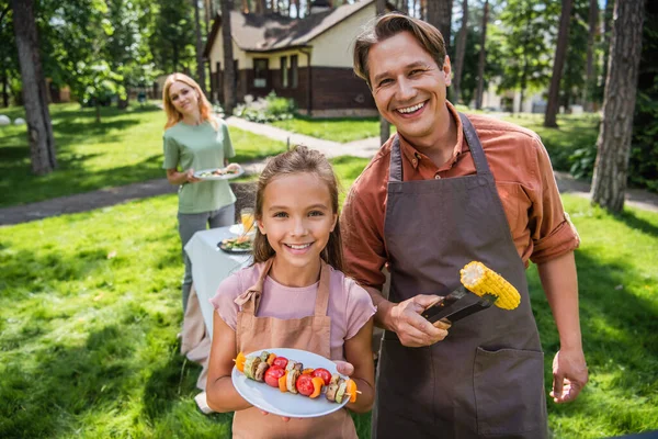Sonriente padre e hijo en delantales sosteniendo verduras a la parrilla al aire libre - foto de stock
