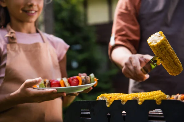 Verschwommene Sicht auf verschwommenen Mann mit gegrilltem Mais in der Nähe von Tochter mit Teller im Freien — Stockfoto