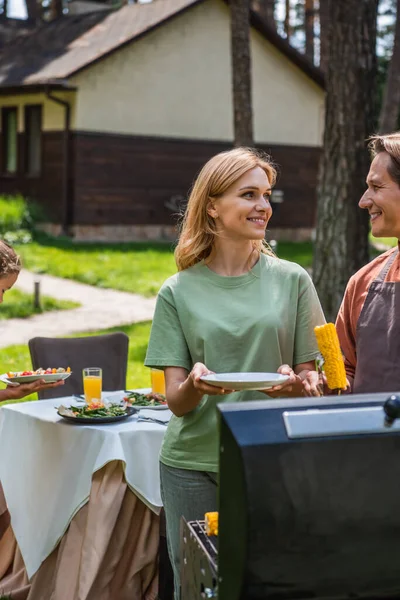 Lächelnder Mann hält Mais neben Grill und Frau mit Teller im Freien — Stockfoto