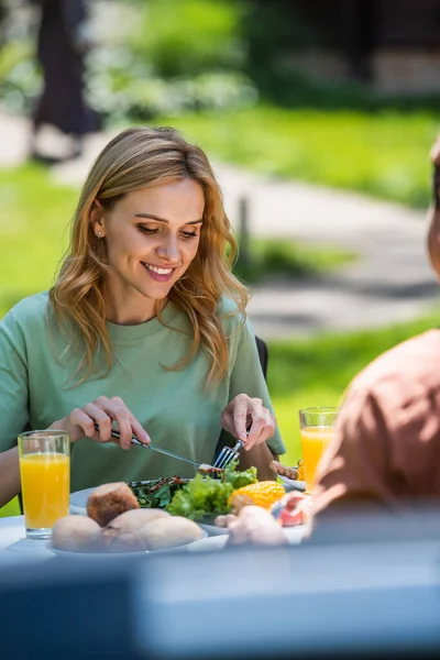 Mujer sonriente sosteniendo cubiertos cerca de la ensalada y esposo borroso al aire libre - foto de stock