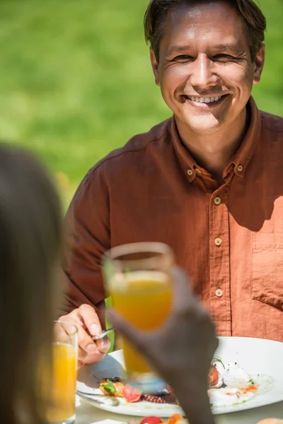 Hombre sonriente mirando a la esposa borrosa con jugo de naranja cerca de la ensalada durante el picnic - foto de stock