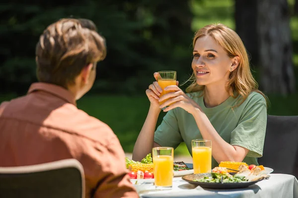 Mulher sorridente com suco de laranja olhando para o marido perto de comida durante o piquenique — Fotografia de Stock