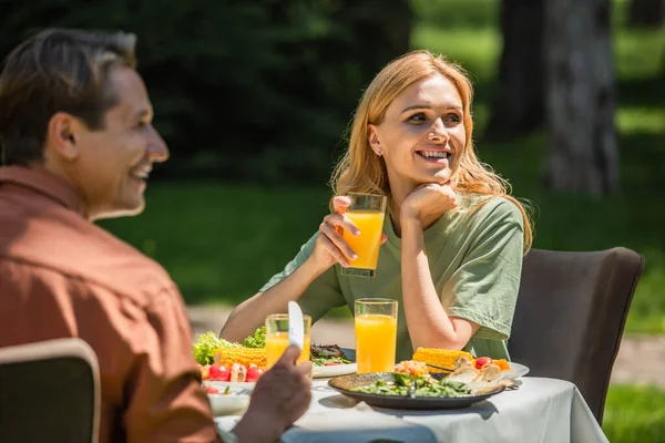 Mujer sonriente con jugo de naranja sentada cerca de marido y comida al aire libre - foto de stock