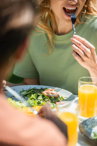 Vista cortada de mulher comendo salada saborosa perto de suco de laranja perto do marido borrado ao ar livre — Fotografia de Stock