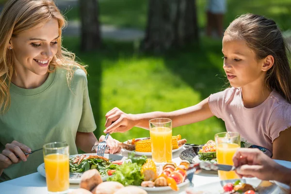 Smiling woman sitting near kid with fork and food during picnic outdoors — Stock Photo