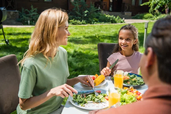 Smiling kid looking at mother with cutlery near food and orange juice outdoors — Stock Photo
