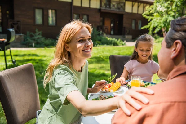 Mujer sonriente tocando marido en primer plano borroso durante el picnic con la familia - foto de stock