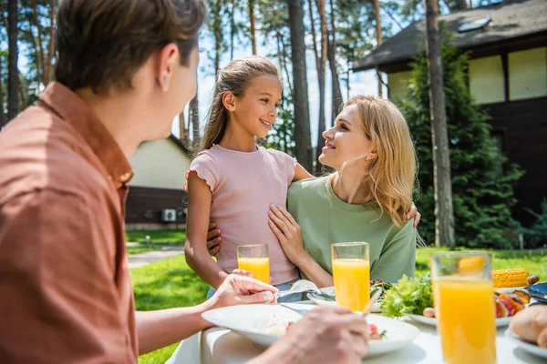 Mujer sonriente abrazando niño cerca borrosa marido y comida al aire libre - foto de stock