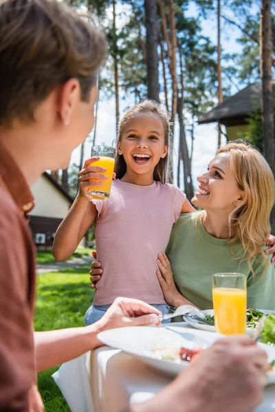 Ragazza sorridente che tiene il succo d'arancia vicino a madre e padre durante il picnic e le vacanze — Foto stock