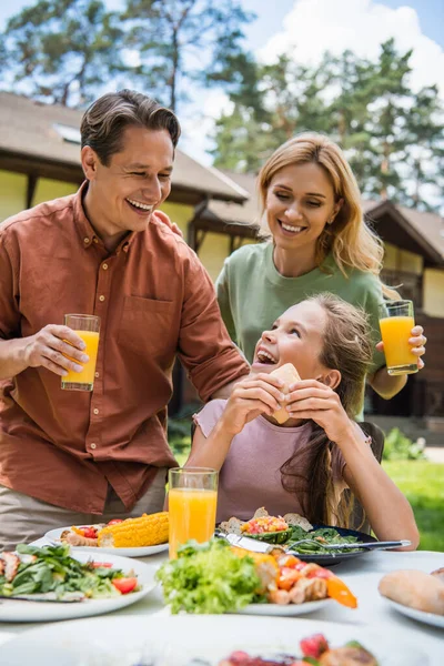 Pais felizes com suco de laranja olhando para a filha perto de comida saborosa durante o piquenique — Fotografia de Stock