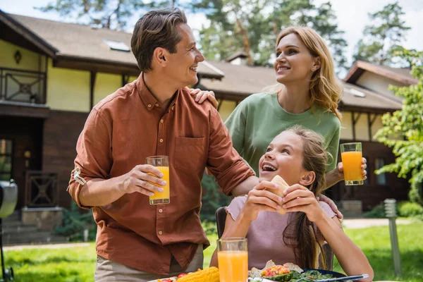 Sonrientes padres con jugo de naranja hablando cerca de un niño con comida durante el fin de semana - foto de stock