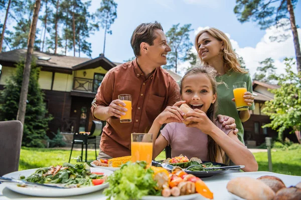 Positives Kind blickt in die Kamera in der Nähe von Essen und Eltern im Freien — Stockfoto