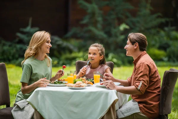 Pais sorridentes falando perto de comida deliciosa e filha ao ar livre — Fotografia de Stock