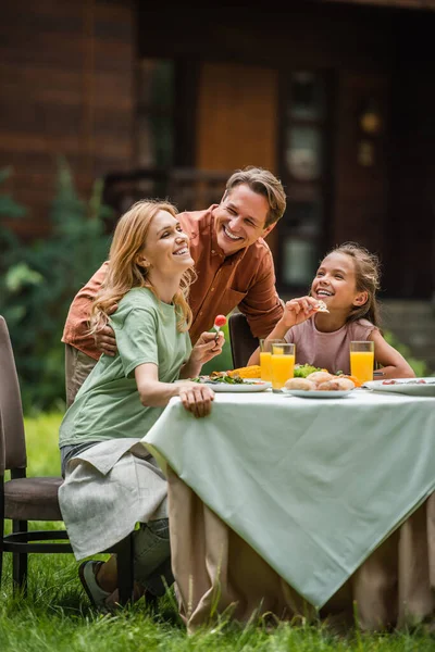 Hombre positivo abrazando esposa e hija durante el picnic en el césped - foto de stock