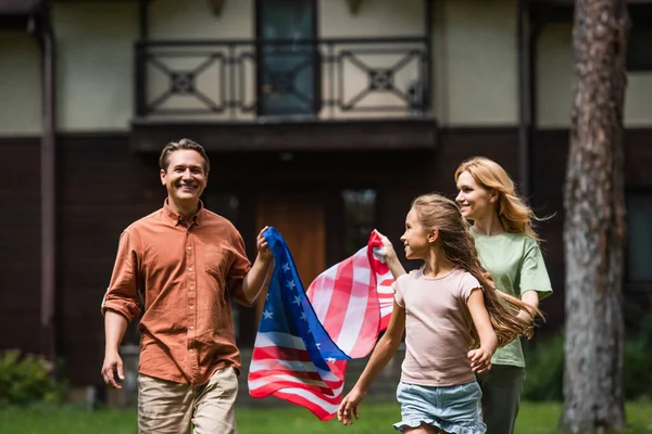 Un homme souriant tenant un drapeau américain près de sa femme et de sa fille à l'extérieur — Photo de stock