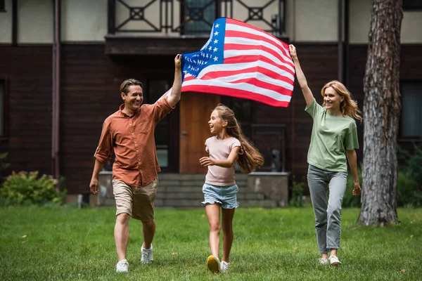 Glückliches Kind spaziert mit amerikanischer Flagge im Freien auf Rasen neben Eltern — Stockfoto