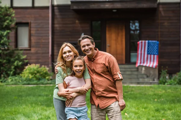 Sonriente familia abrazándose cerca borrosa bandera americana en casa al aire libre - foto de stock