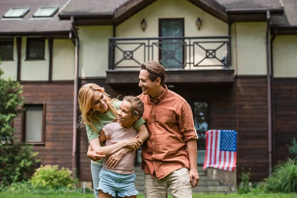 Sonriente familia abrazando hija cerca borrosa bandera americana en casa de vacaciones - foto de stock