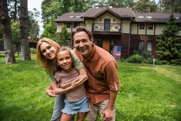 Hombre positivo mirando a la cámara mientras la esposa abrazando a la hija al aire libre - foto de stock